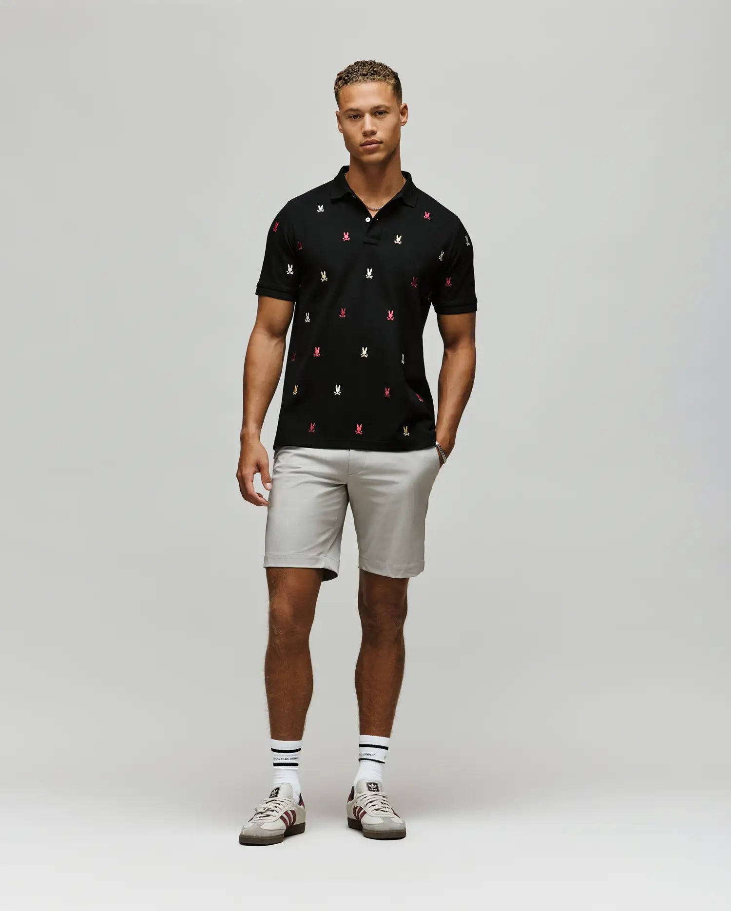A man stands against a light gray background wearing the Psycho Bunny MENS HOLLAND ALL OVER BUNNY PIQUE POLO (B6K618F200), paired with light gray shorts, striped socks, and white sneakers. His hands rest relaxed at his sides.