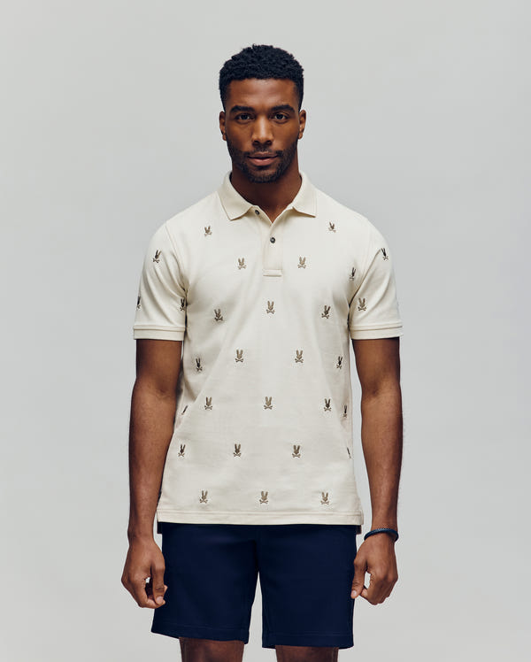 A man faces forward in a Psycho Bunny MENS HOLLAND ALL OVER BUNNY PIQUE POLO (B6K618F200), featuring a light color and repeated bunny logo pattern, paired with dark shorts, against a plain light gray background.