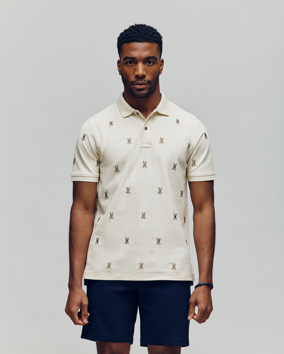 A man faces forward in a Psycho Bunny MENS HOLLAND ALL OVER BUNNY PIQUE POLO (B6K618F200), featuring a light color and repeated bunny logo pattern, paired with dark shorts, against a plain light gray background.