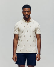 A man faces forward in a Psycho Bunny MENS HOLLAND ALL OVER BUNNY PIQUE POLO (B6K618F200), featuring a light color and repeated bunny logo pattern, paired with dark shorts, against a plain light gray background.
