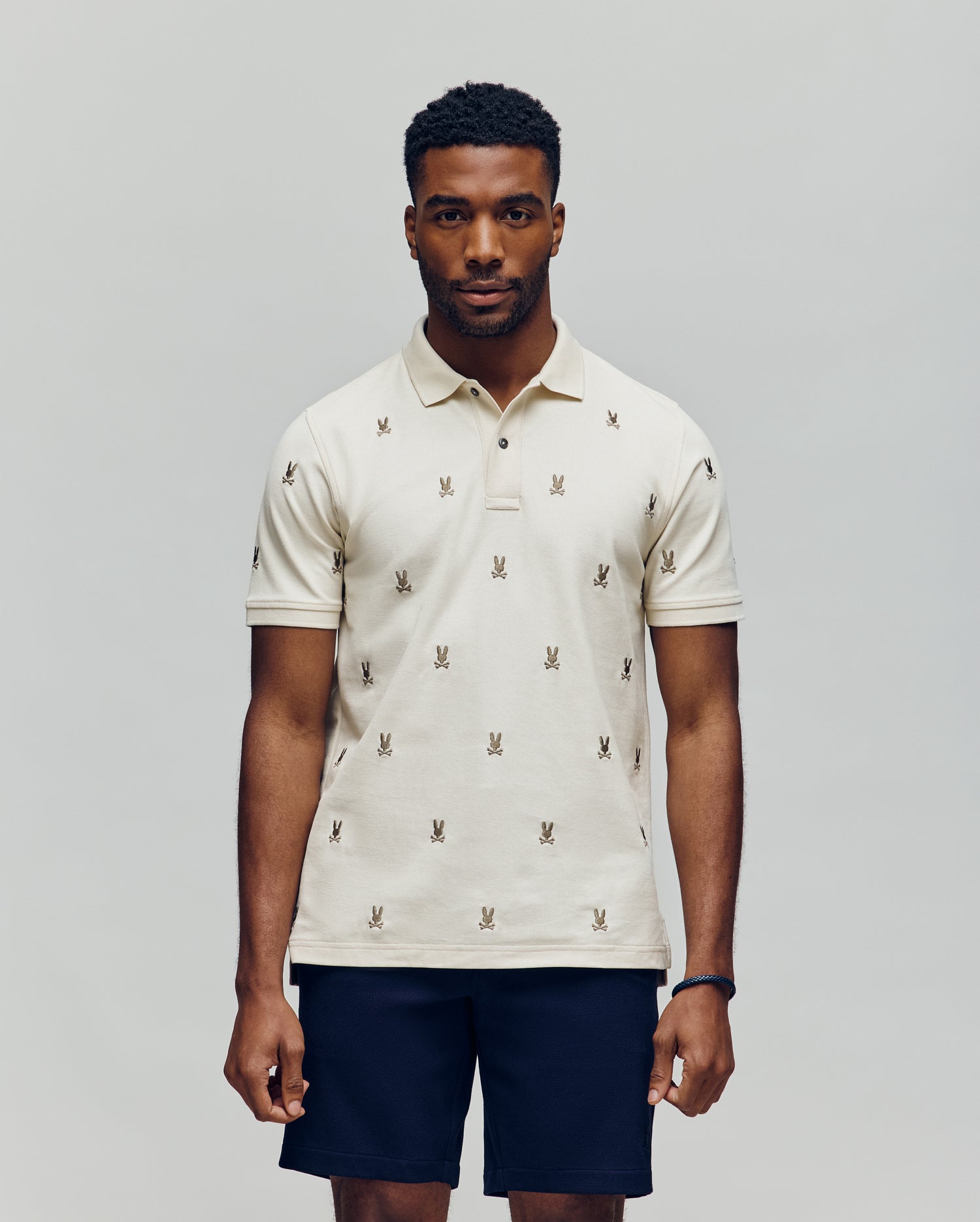 A man faces forward in a Psycho Bunny MENS HOLLAND ALL OVER BUNNY PIQUE POLO (B6K618F200), featuring a light color and repeated bunny logo pattern, paired with dark shorts, against a plain light gray background.