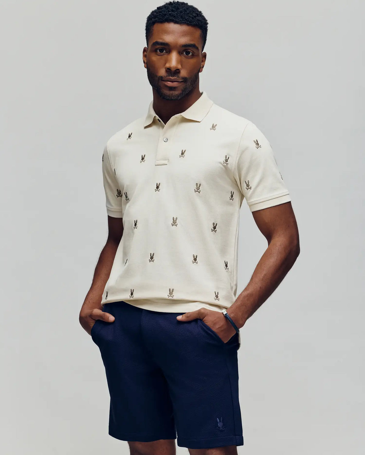 A man stands with his hands in his pockets, wearing the Psycho Bunny MENS HOLLAND ALL OVER BUNNY PIQUE POLO (B6K618F200) in cream with embroidered logos and navy blue shorts, set against a plain light background.