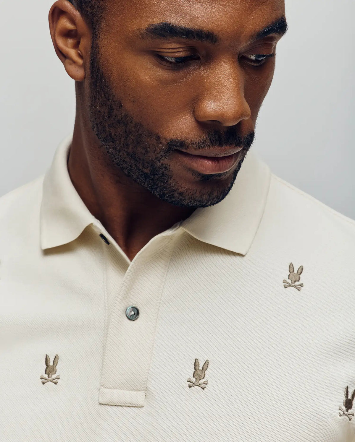 A man wearing the Psycho Bunny MENS HOLLAND ALL OVER BUNNY PIQUE POLO (B6K618F200), featuring embroidered bunny skull-and-crossbones motifs, looks down with his upper torso and face visible against a plain background.