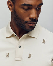 A man wearing the Psycho Bunny MENS HOLLAND ALL OVER BUNNY PIQUE POLO (B6K618F200), featuring embroidered bunny skull-and-crossbones motifs, looks down with his upper torso and face visible against a plain background.