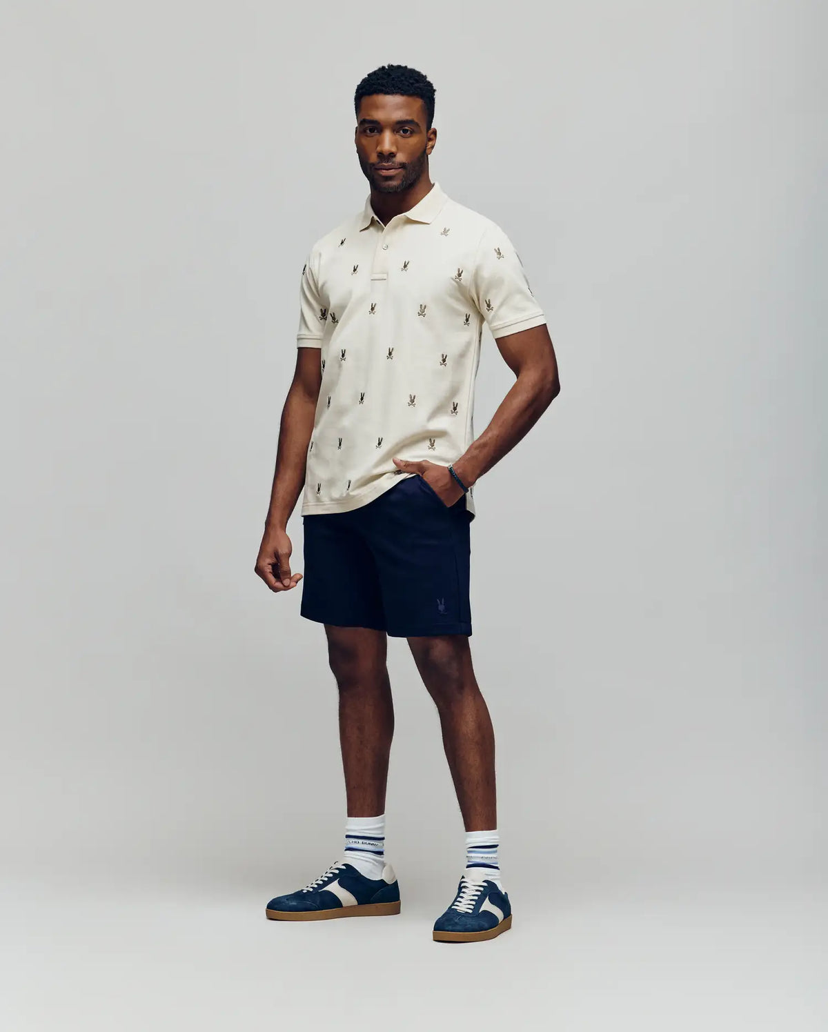 A man stands against a plain background wearing the Psycho Bunny MENS HOLLAND ALL OVER BUNNY PIQUE POLO (B6K618F200), dark navy shorts, striped socks, and blue sneakers. One hand is in his pocket as he looks at the camera.