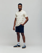A man stands against a plain background wearing the Psycho Bunny MENS HOLLAND ALL OVER BUNNY PIQUE POLO (B6K618F200), dark navy shorts, striped socks, and blue sneakers. One hand is in his pocket as he looks at the camera.