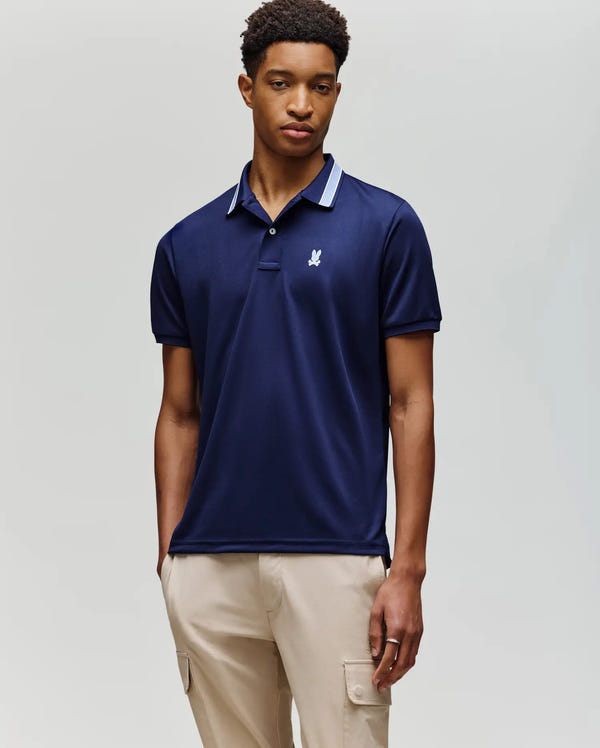 A young man wears the Psycho Bunny MENS FORREST SPORT PIQUE POLO (B6K660F200) in navy with a white emblem and striped collar, paired with light beige cargo pants, against a plain light background.