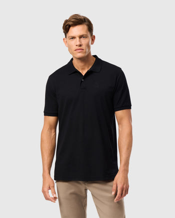A person dressed in a Psycho Bunny MENS JAMES PREMIUM PIQUE POLO SHIRT in black, paired with beige pants, stands against a plain light gray background, exemplifying timeless style with comfort.