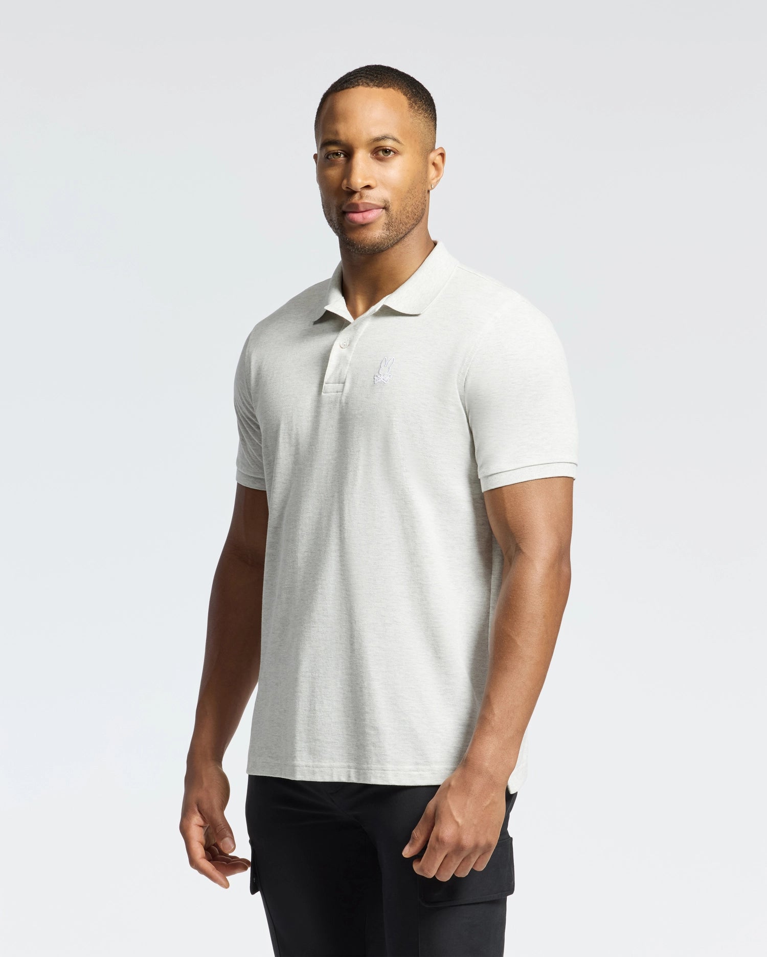 A person in a slim-fit Mens James Pique Polo (B6K945D200) by Psycho Bunny, made from durable light gray Pima cotton, paired with black pants, stands against a white background. They hold a neutral expression while gazing slightly to the side.