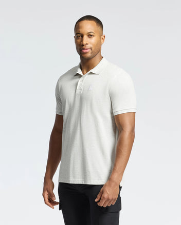 A person in a slim-fit Mens James Pique Polo (B6K945D200) by Psycho Bunny, made from durable light gray Pima cotton, paired with black pants, stands against a white background. They hold a neutral expression while gazing slightly to the side.