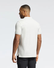 A man with short hair is turned away, highlighting the slim fit MENS JAMES PIQUE POLO - B6K945D200 in light gray by Psycho Bunny, made of durable Pima cotton, paired with black pants against a plain white background.