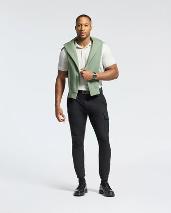 A man stands casually in a light gray Psycho Bunny MENS JAMES PIQUE POLO - B6K945D200, made of durable Pima cotton, paired with black pants and shoes. A green sweatshirt is draped over his shoulders, and he wears a wristwatch against a plain white background.