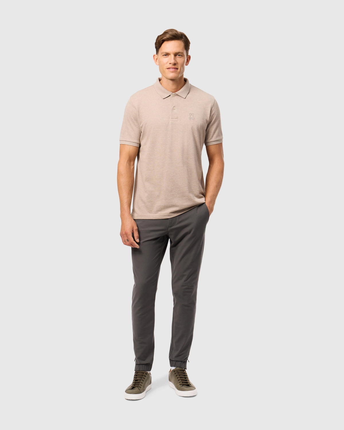Against a plain backdrop, a man dons the Psycho Bunny MENS JAMES PREMIUM PIQUE POLO SHIRT, dark gray pants, and olive green sneakers. The Pima cotton shirt exudes relaxed elegance as he poses with one hand in his pocket.