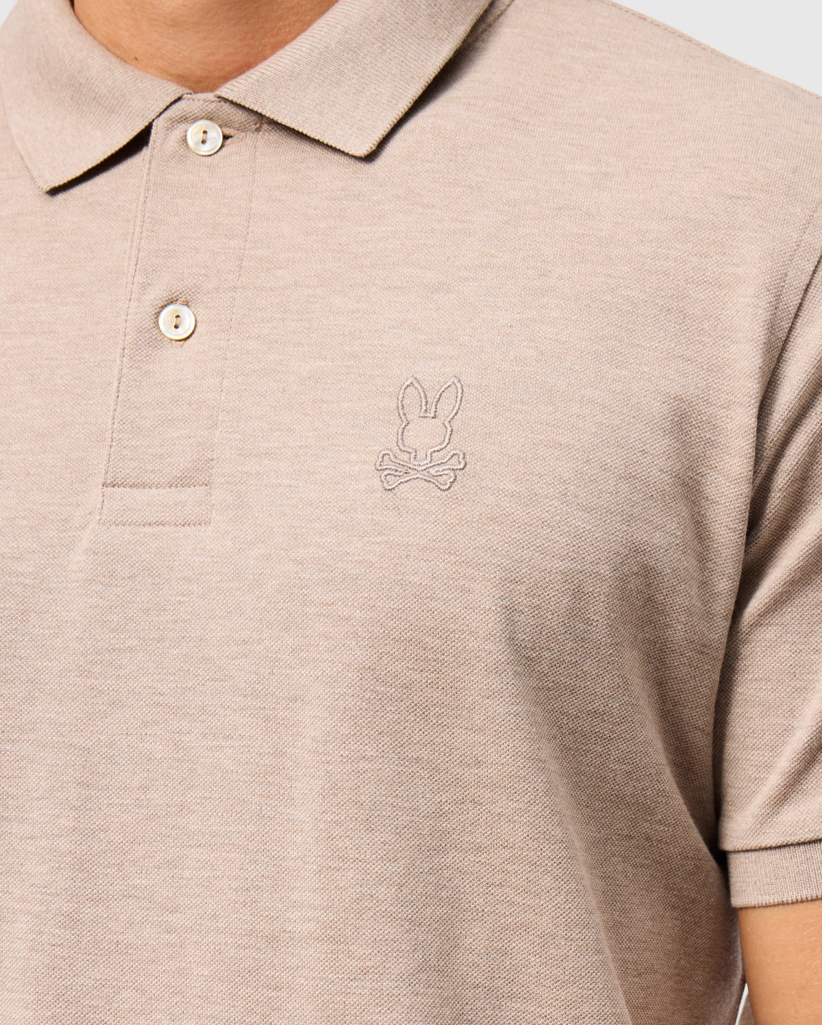 A person is wearing the MENS JAMES PREMIUM PIQUE POLO SHIRT - B6K945D200 by Psycho Bunny, made from soft Pima cotton. This beige polo features a bunny and crossbones logo embroidered on the left chest, with a collar and two buttons for style and durability.