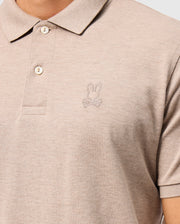 A person is wearing the MENS JAMES PREMIUM PIQUE POLO SHIRT - B6K945D200 by Psycho Bunny, made from soft Pima cotton. This beige polo features a bunny and crossbones logo embroidered on the left chest, with a collar and two buttons for style and durability.