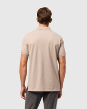 A man with short brown hair wears a Psycho Bunny MENS JAMES PREMIUM PIQUE POLO SHIRT in beige and dark gray pants, standing with his back to the camera against a light gray background.