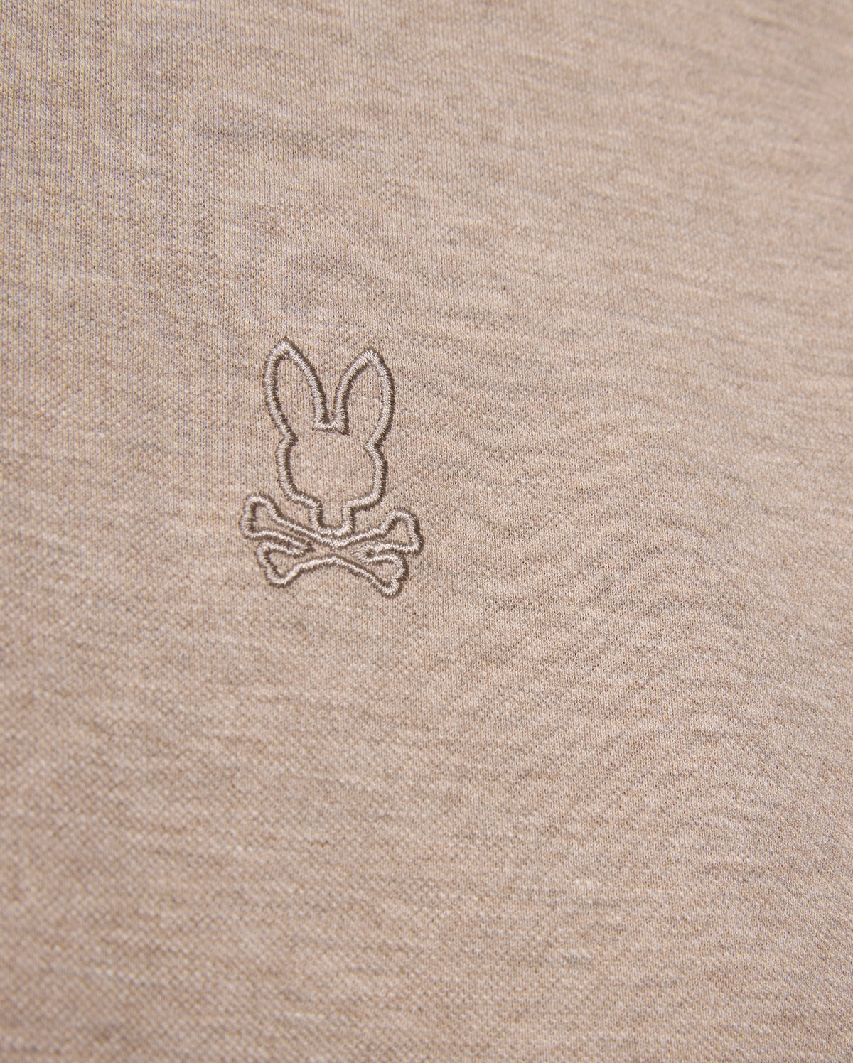 A close-up of a beige Pima cotton fabric with a stitched emblem showing a bunny head above two crossed bones, perfect for a stylish and comfortable MENS JAMES PREMIUM PIQUE POLO SHIRT - B6K945D200 by Psycho Bunny.