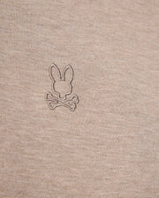 A close-up of a beige Pima cotton fabric with a stitched emblem showing a bunny head above two crossed bones, perfect for a stylish and comfortable MENS JAMES PREMIUM PIQUE POLO SHIRT - B6K945D200 by Psycho Bunny.
