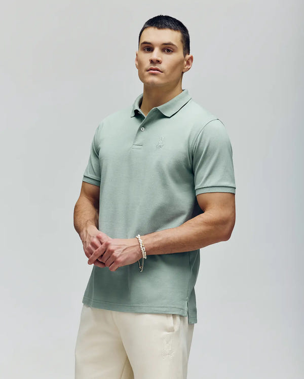 A man with short dark hair wears the Psycho Bunny MENS JAMES PREMIUM PIQUE POLO (B6K945D200) in light green, featuring a crease-resistant collar, styled with cream-colored pants against a plain light background. He's looking slightly off-camera.