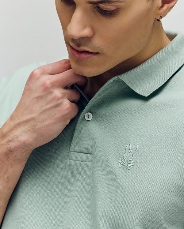A man wearing the Psycho Bunny MENS JAMES PREMIUM PIQUE POLO (B6K945D200) in light green, featuring the signature bunny and crossbones logo, looks down while holding the collar of his durable Pima cotton shirt.