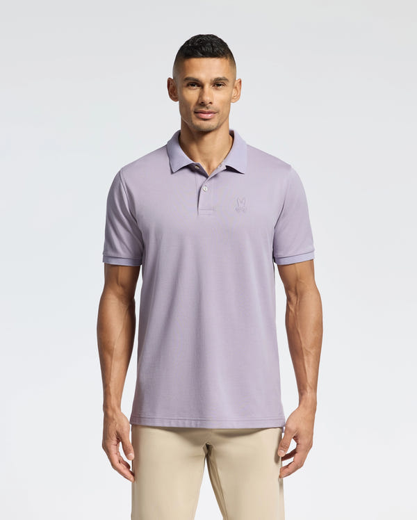A person stands against a plain white background wearing the Psycho Bunny MENS JAMES PIQUE POLO - B6K945D200 in light purple, paired with beige pants. The durable Pima cotton shirt has a subtle logo on the chest and a crease-resistant collar.
