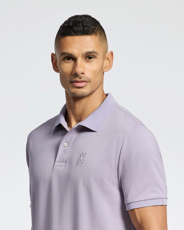 Against a plain background, someone with short hair wears the Psycho Bunny MENS JAMES PIQUE POLO in light purple. The shirt, B6K945D200, features a small embroidered rabbit logo on the left and has a crease-resistant collar. Their expression is neutral.