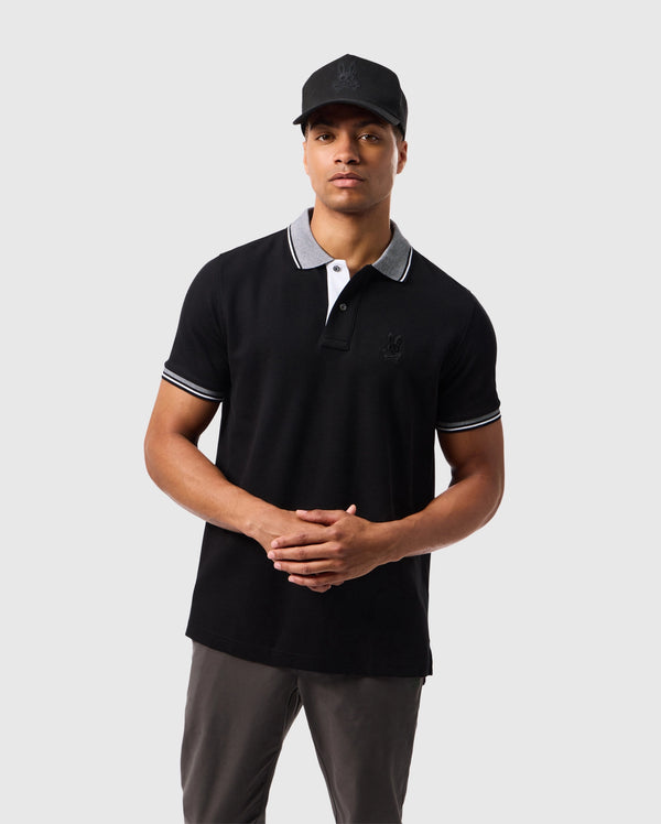 A man dressed in the Psycho Bunny Men's Malton Pique Polo Shirt (B6K950D200) in black with white trim on the collar and sleeves, paired with grey pants, stands with his hands clasped together. The high-quality Pima cotton fabric of the polo adds a touch of sophistication. He is also wearing a black baseball cap. The background is a plain light grey.