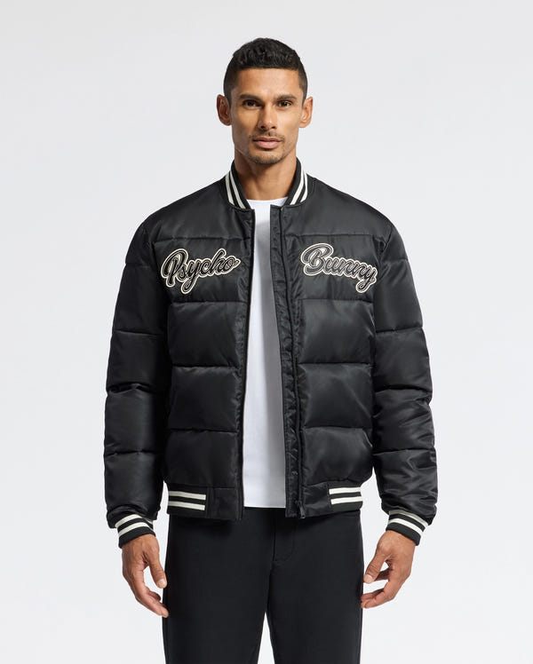 A man confidently wears the MENS FRAZER FASHION PUFFER - B6N338E200 from Psycho Bunny, showcasing its black design with "Psycho Bunny" embroidered on the chest. This fashionable piece is accented with white stripes on the collar, cuffs, and hem as he poses against a plain white background.