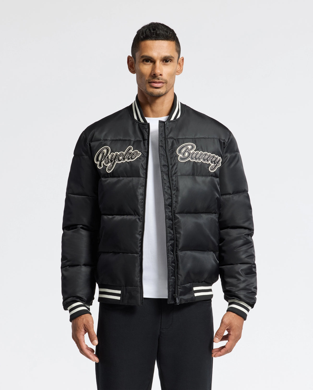 A man confidently wears the MENS FRAZER FASHION PUFFER - B6N338E200 from Psycho Bunny, showcasing its black design with "Psycho Bunny" embroidered on the chest. This fashionable piece is accented with white stripes on the collar, cuffs, and hem as he poses against a plain white background.