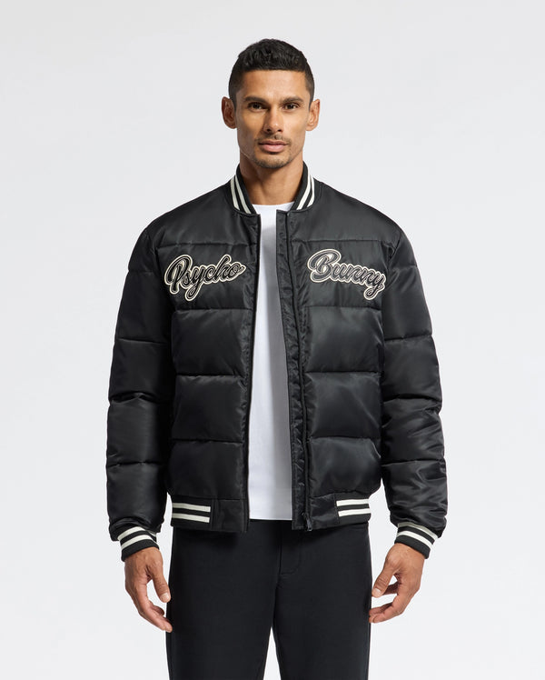 A man confidently wears the MENS FRAZER FASHION PUFFER - B6N338E200 from Psycho Bunny, showcasing its black design with "Psycho Bunny" embroidered on the chest. This fashionable piece is accented with white stripes on the collar, cuffs, and hem as he poses against a plain white background.