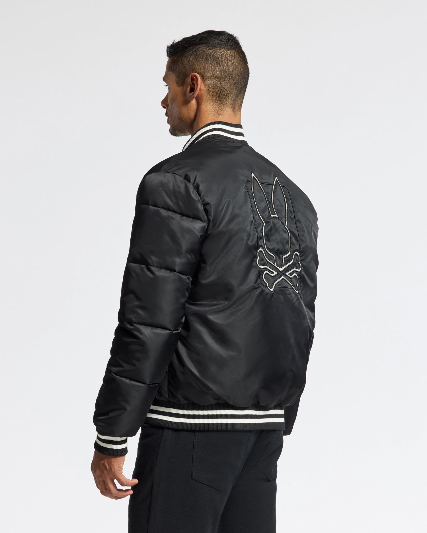 A man is seen facing away from the camera, highlighting the stylish MENS FRAZER FASHION PUFFER - B6N338E200 by Psycho Bunny. The black bomber jacket features a white-outlined rabbit and crossbones design on the back, embodying the edgy side of men's fashion jackets as he stands against a plain white background.