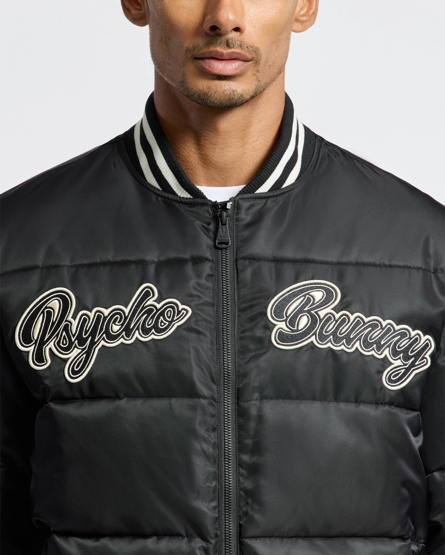 A person wearing the MENS FRAZER FASHION PUFFER - B6N338E200 by Psycho Bunny. The jacket features a ribbed collar with white stripes and a zipper closure, perfectly blending style and comfort.