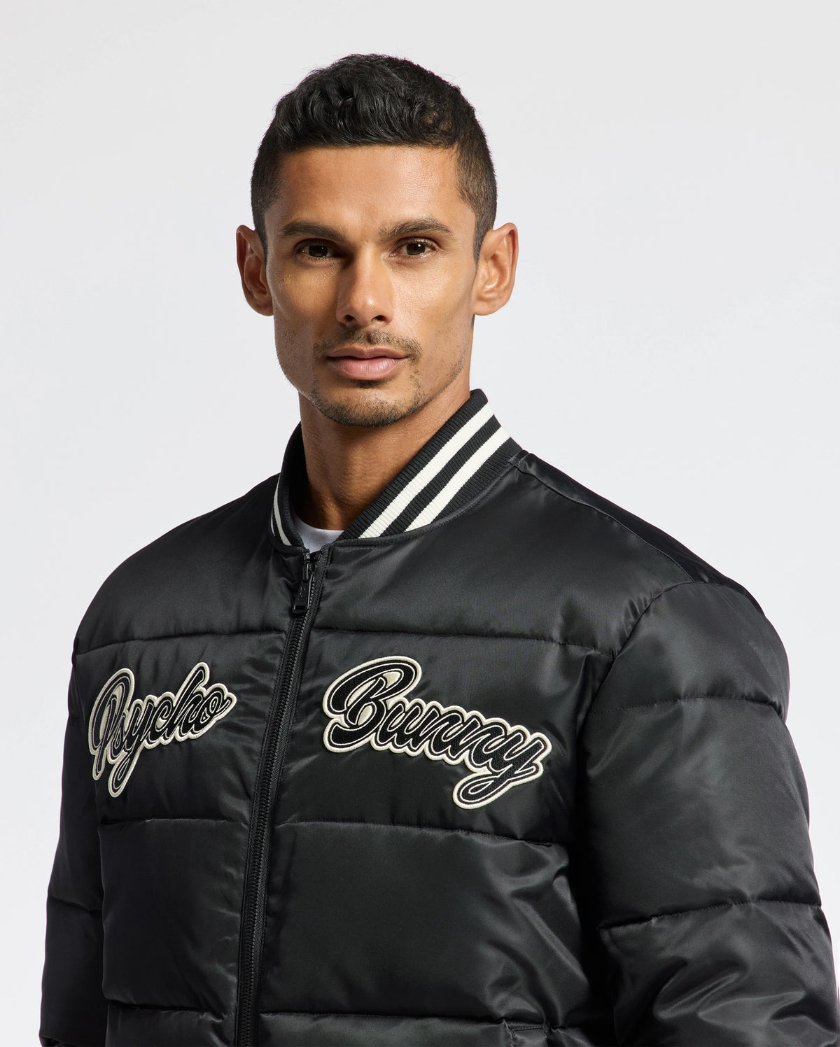 A person wearing the Psycho Bunny MENS FRAZER FASHION PUFFER - B6N338E200, which has embroidered text, stands in front of a plain white background. The stylish jacket features a striped collar, embodying the latest trends in men's fashion jackets.