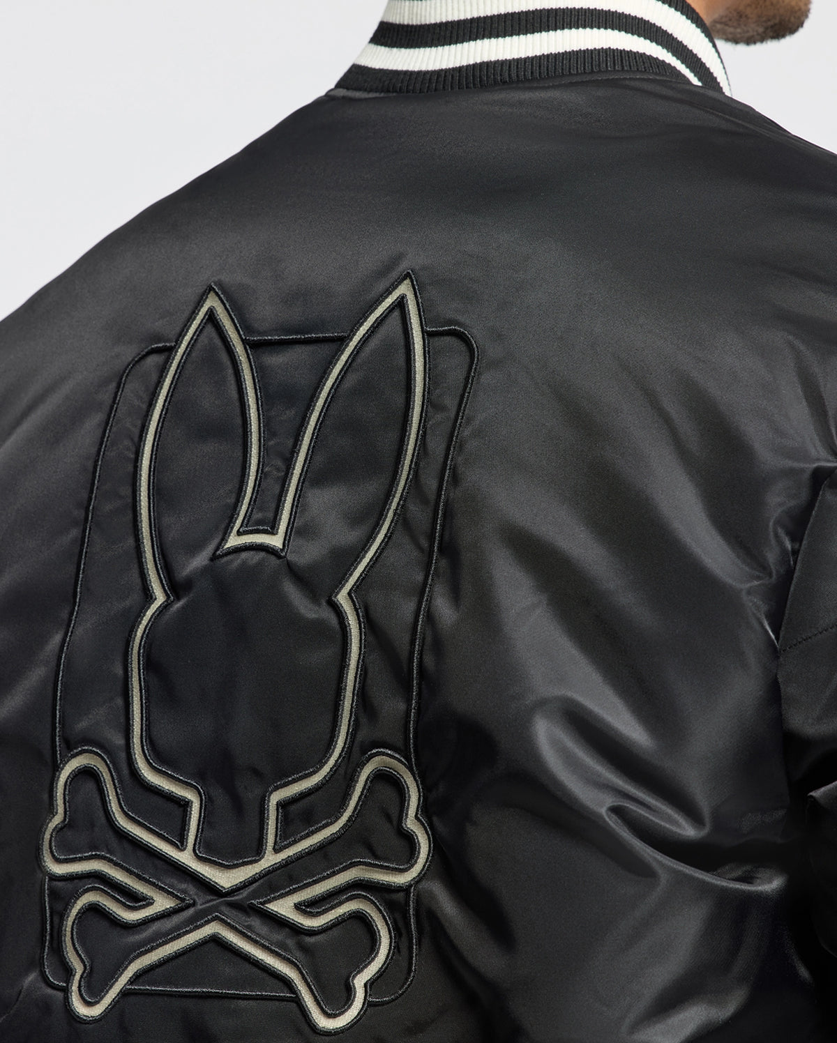 A close-up of an individual wearing the Psycho Bunny Men's Frazer Fashion Puffer - B6N338E200 showcases a black bomber-style jacket. This stylish piece features an embroidered bunny skull and crossbones on the back, paired with a sleek striped collar.
