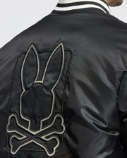 A close-up of an individual wearing the Psycho Bunny Men's Frazer Fashion Puffer - B6N338E200 showcases a black bomber-style jacket. This stylish piece features an embroidered bunny skull and crossbones on the back, paired with a sleek striped collar.