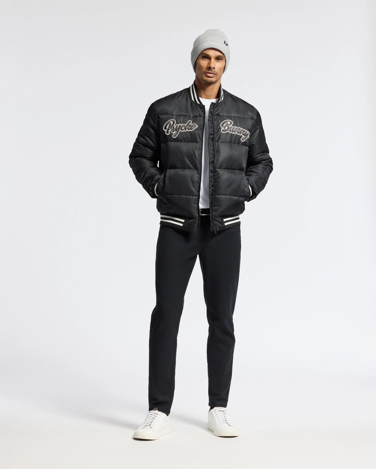 A man wearing a Psycho Bunny MENS FRAZER FASHION PUFFER - B6N338E200 stands against a plain white background. Hands in his pockets, he looks confidently at the camera, showcasing his stylish ensemble comprising a gray beanie, black varsity jacket, and white shirt with ease.