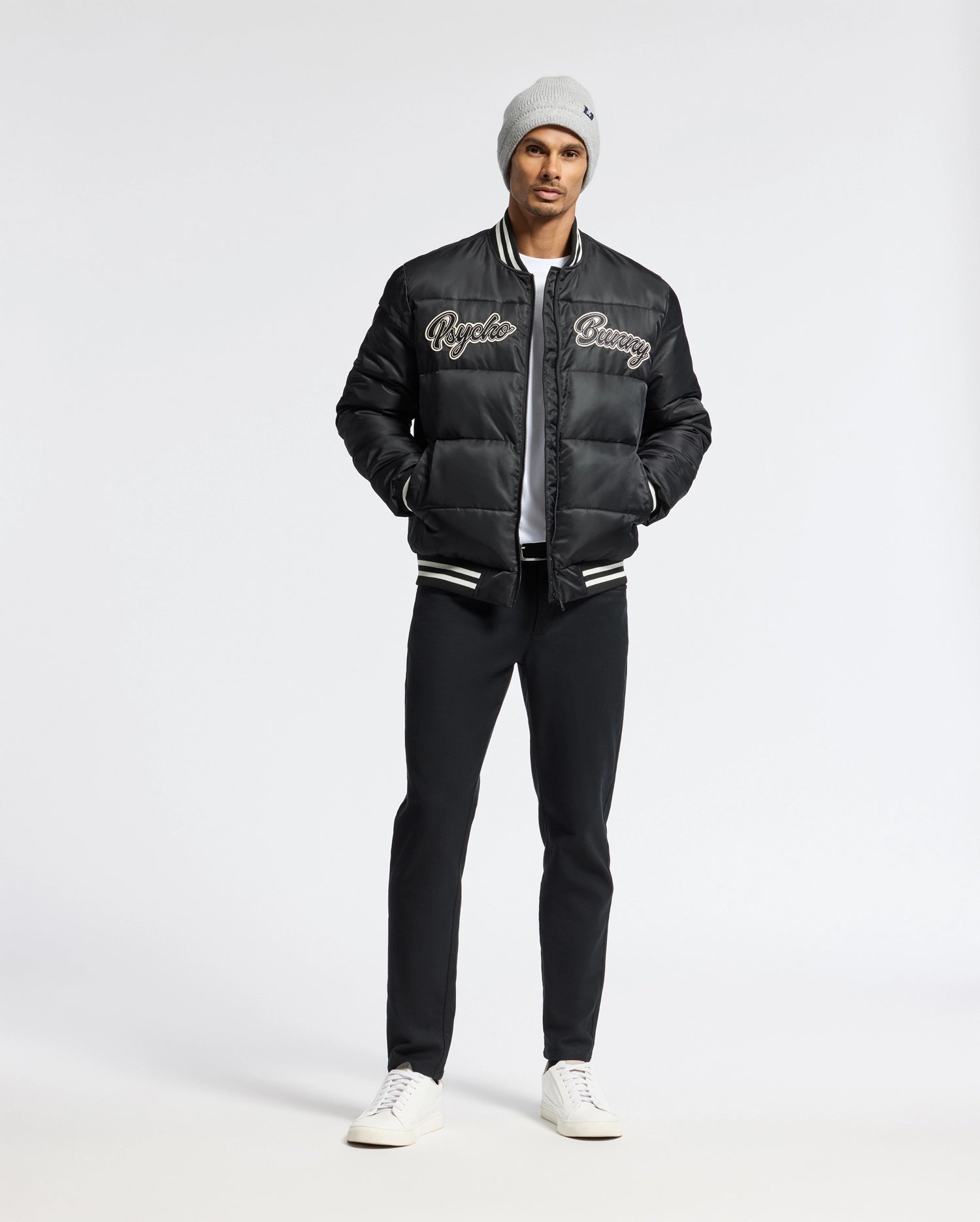 A man wearing a Psycho Bunny MENS FRAZER FASHION PUFFER - B6N338E200 stands against a plain white background. Hands in his pockets, he looks confidently at the camera, showcasing his stylish ensemble comprising a gray beanie, black varsity jacket, and white shirt with ease.