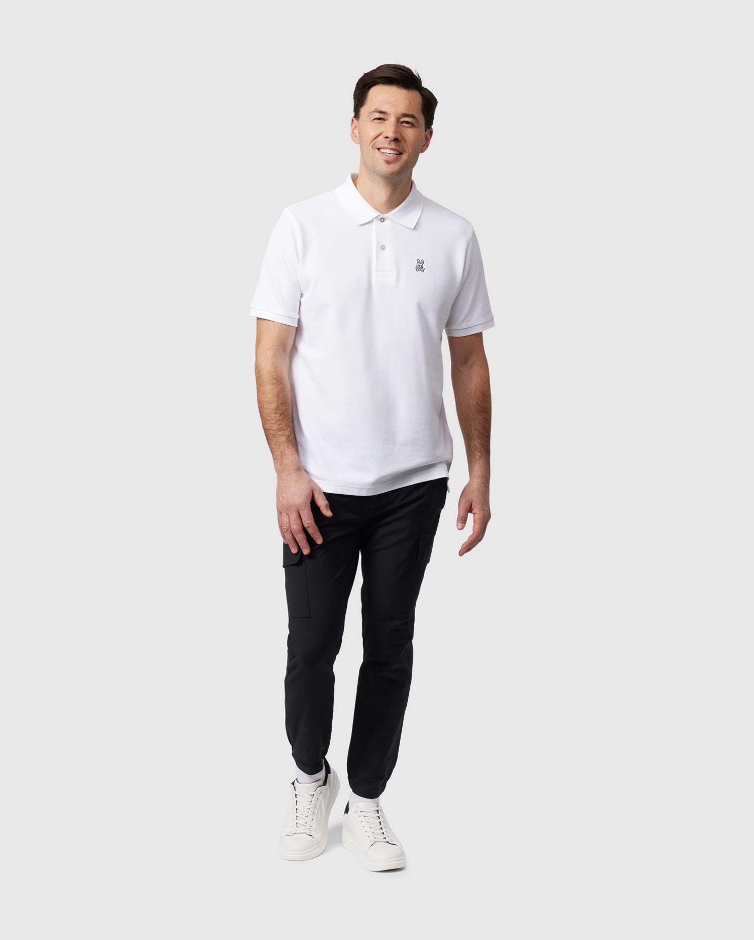 A man is standing against a plain white background, wearing a white polo shirt, Psycho Bunny MENS LLOYD CARGO JOGGER - B6P159B200 with utilitarian pockets, and white sneakers. He is smiling and has one hand slightly raised in front of him while the other rests by his side.