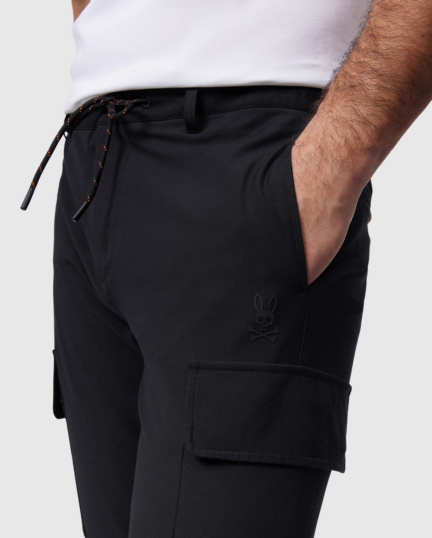 A person wearing the Psycho Bunny MENS LLOYD CARGO JOGGER - B6P159B200, featuring black cargo jogger pants with a drawstring waist and a subtle rabbit skull logo on the pocket. The pants have utilitarian pockets, including a large one on the thigh, and elasticized cuffs. The person’s hand is in one pocket, and they are wearing a white shirt.