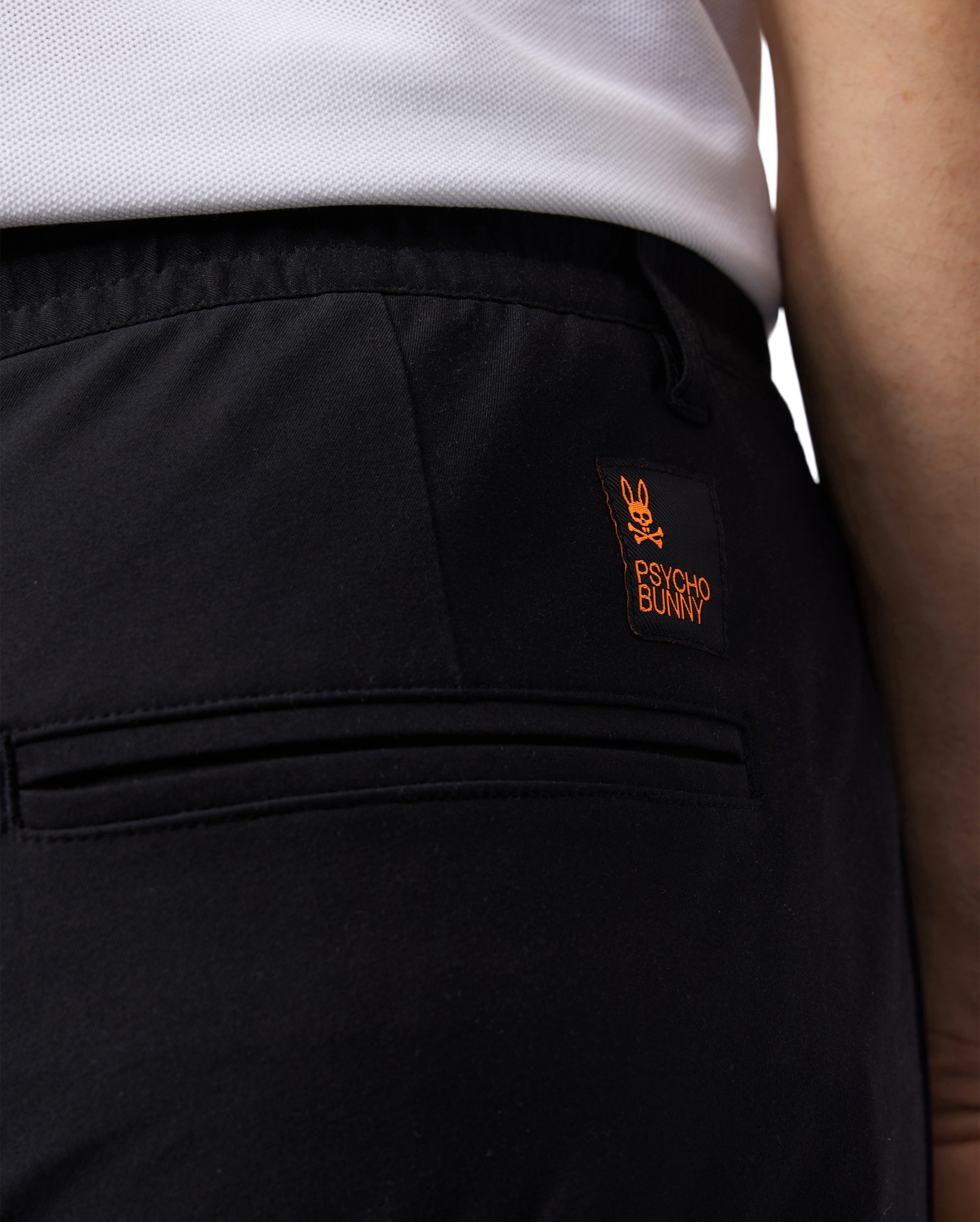 Close-up of the back of a person's pants. The MENS LLOYD CARGO JOGGER - B6P159B200 by Psycho Bunny are black joggers with a small orange and black tag featuring the brand's bunny logo and the words "Psycho Bunny" sewn above one of the utilitarian pockets. The person is wearing a white shirt tucked into the pants, which also feature elasticized cuffs.