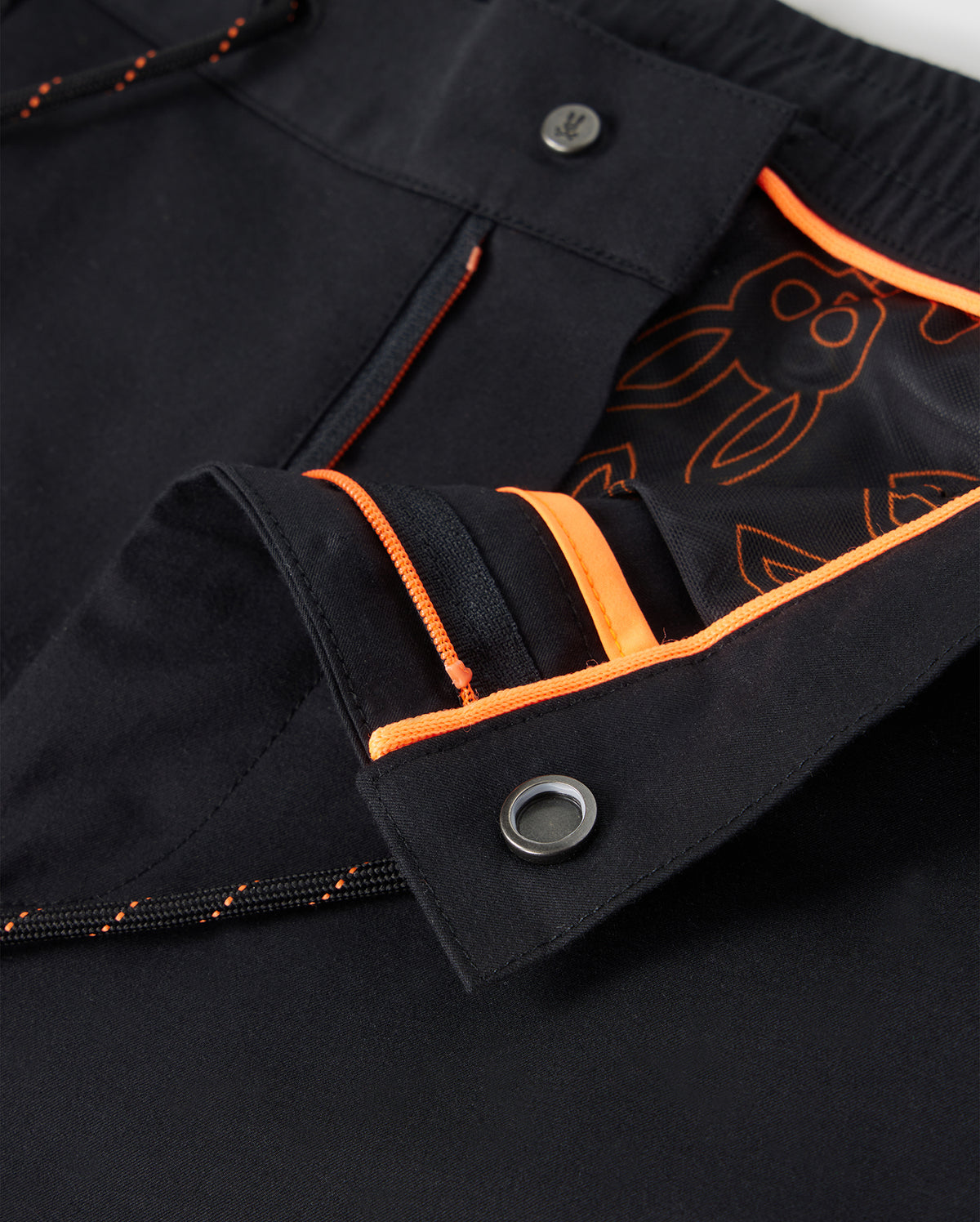 Close-up of the MENS LLOYD CARGO JOGGER - B6P159B200 by Psycho Bunny, featuring black cargo jogger pants with orange stitching and an orange-lined waistband. The interior fabric showcases an orange design with cartoon characters. These pants are designed with two silver buttons, a zippered opening equipped with snap closures, and elasticized cuffs for a snug fit.