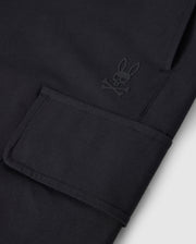 Close-up image of a black fabric with a pocket flap in focus. Adjacent to the pocket is a subtle, embossed logo of Psycho Bunny's bunny head with crossbones beneath it, adding a unique design detail to the material. The utilitarian pockets add both functionality and style to these MENS LLOYD CARGO JOGGER - B6P159B200 by Psycho Bunny.