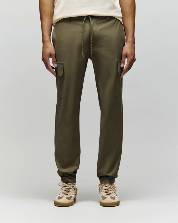 A person wears Psycho Bunny MENS LLOYD CARGO PANT - B6P159B200, olive green joggers with side pockets and drawstring waist, paired with beige and tan sneakers, standing against a plain light background.