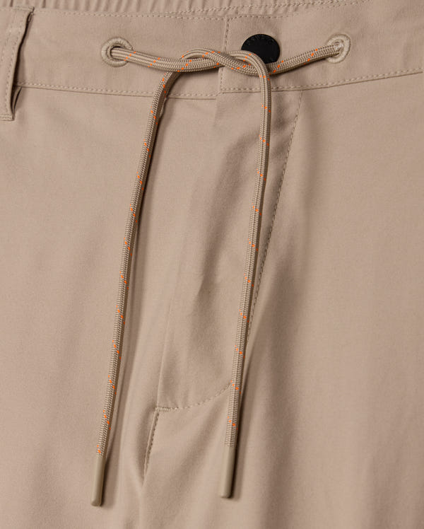 Close-up image of the MENS LLOYD CARGO JOGGER - B6P159B200 by Psycho Bunny, showcasing the beige jogger pants with a black button at the waistband, an elastic drawstring with orange and white stripes for tightening, and a side pocket. The fabric appears smooth and slightly matte, exuding subtle embroidery refinement.