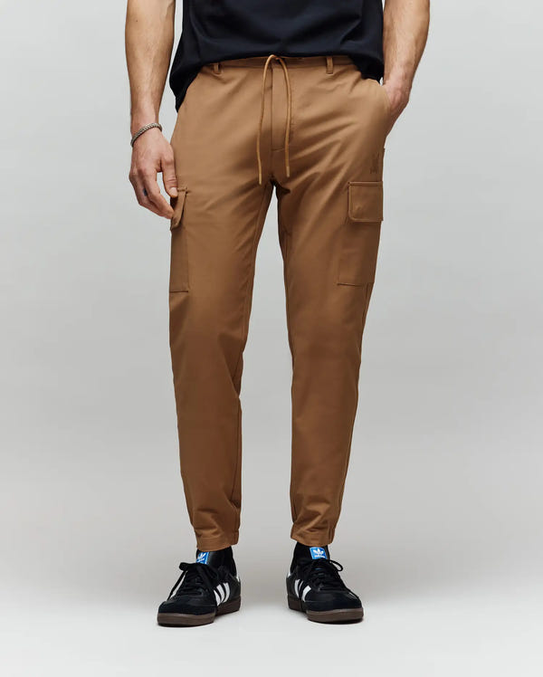 A person wearing Psycho Bunny MENS LLOYD CARGO PANT - B6P159B200 in tan, paired with a black t-shirt and black Adidas sneakers with white stripes, is posed against a plain light grey background.