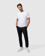 A man in a white t-shirt and Psycho Bunny MENS MADISON COMMUTER PANT - B6P395Z1WB stands casually with one hand in his pocket, on a plain light gray background. He wears white sneakers and looks directly at the camera with a soft gaze.