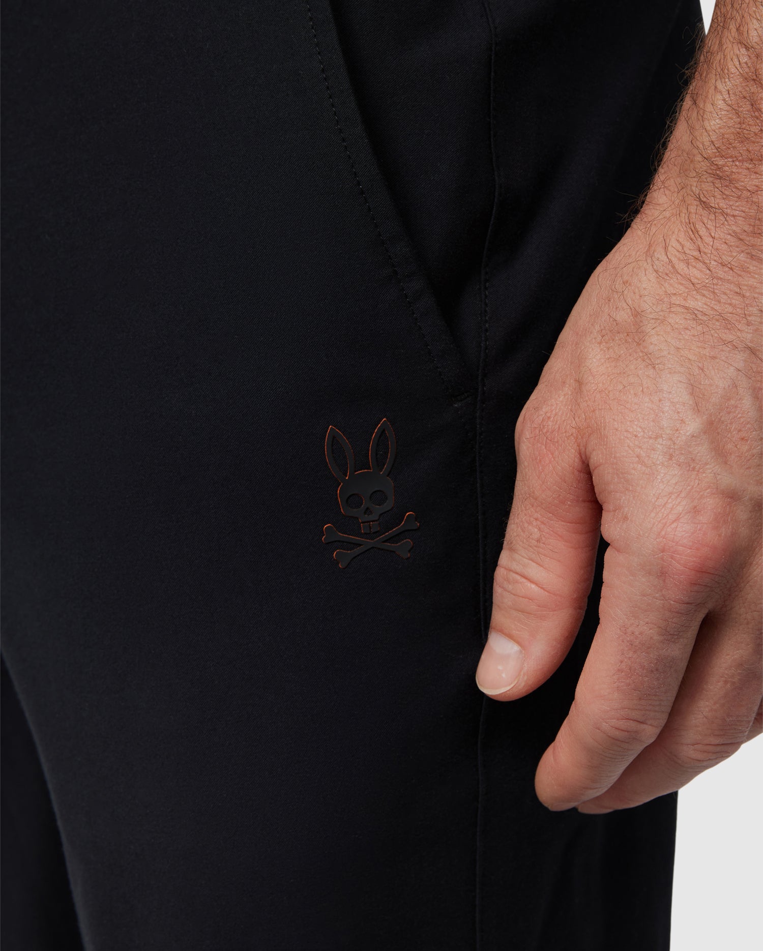 Close-up of a black Men's Madison Commuter Pant by Psycho Bunny made from stretch woven material with a small, dark embroidered logo featuring a stylized rabbit with crossed bones, located near a person's hand.