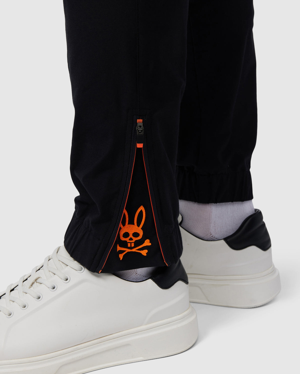 A close-up of a person's lower legs wearing Psycho Bunny's MENS MADISON COMMUTER PANT - B6P395Z1WB, with a zipper at the ankle, revealing an orange rabbit design on a gray inner lining, paired with white shoes.