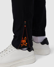 A close-up of a person's lower legs wearing Psycho Bunny's MENS MADISON COMMUTER PANT - B6P395Z1WB, with a zipper at the ankle, revealing an orange rabbit design on a gray inner lining, paired with white shoes.