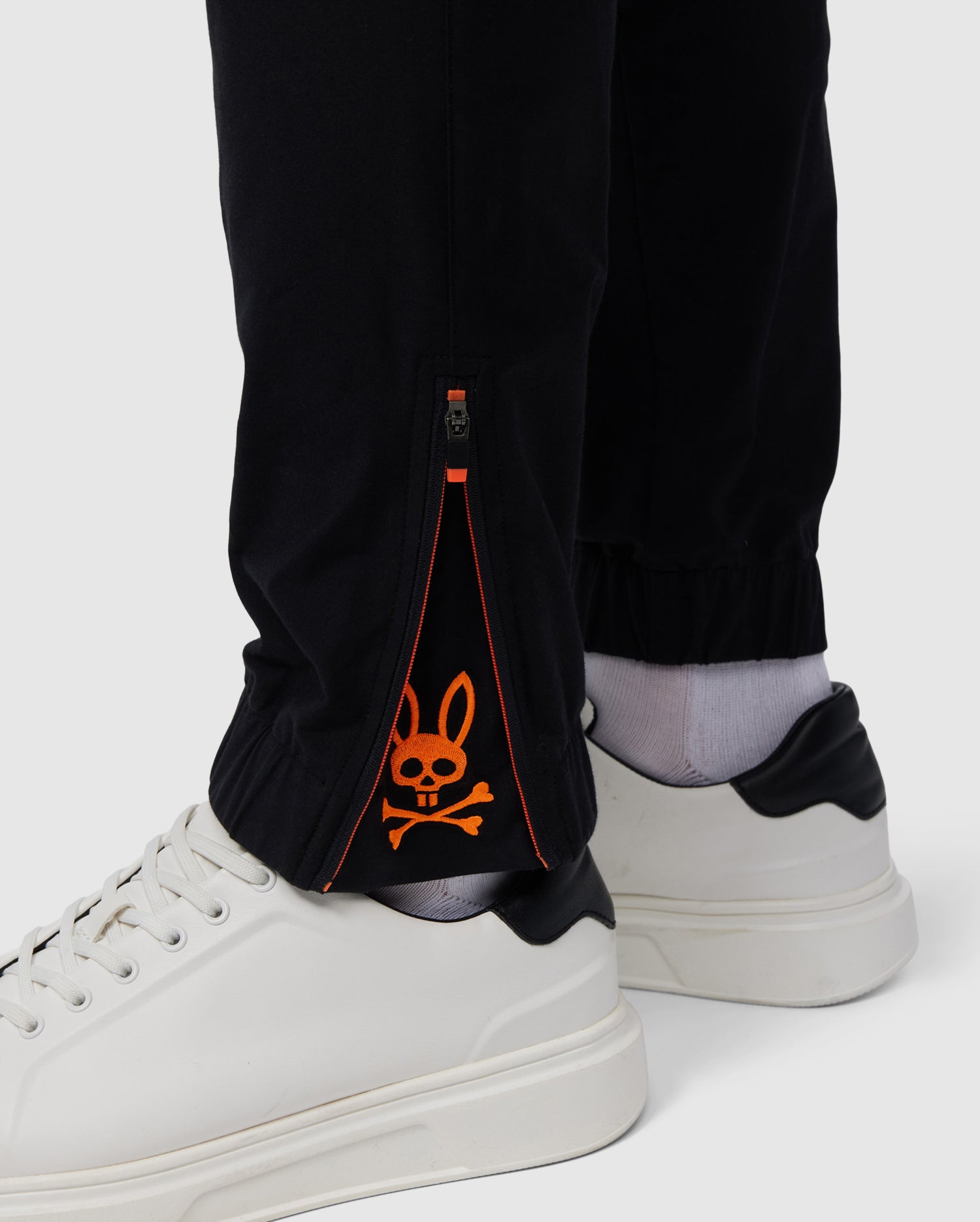 A close-up of a person's lower legs wearing Psycho Bunny's MENS MADISON COMMUTER PANT - B6P395Z1WB, with a zipper at the ankle, revealing an orange rabbit design on a gray inner lining, paired with white shoes.